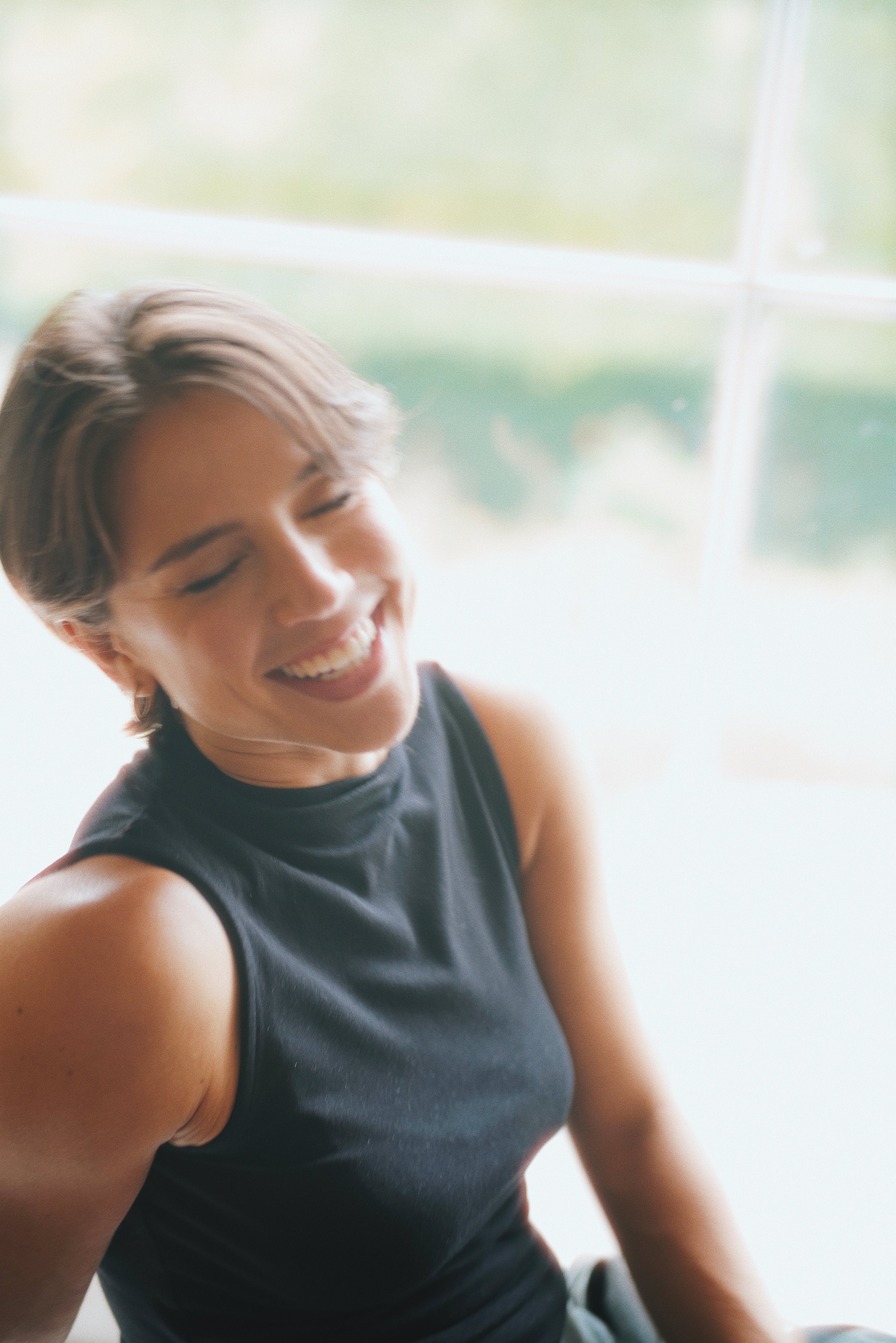 Close-up of a smiling woman with short, light brown hair, wearing a sleeveless black top, sitting indoors with a large window in the background.