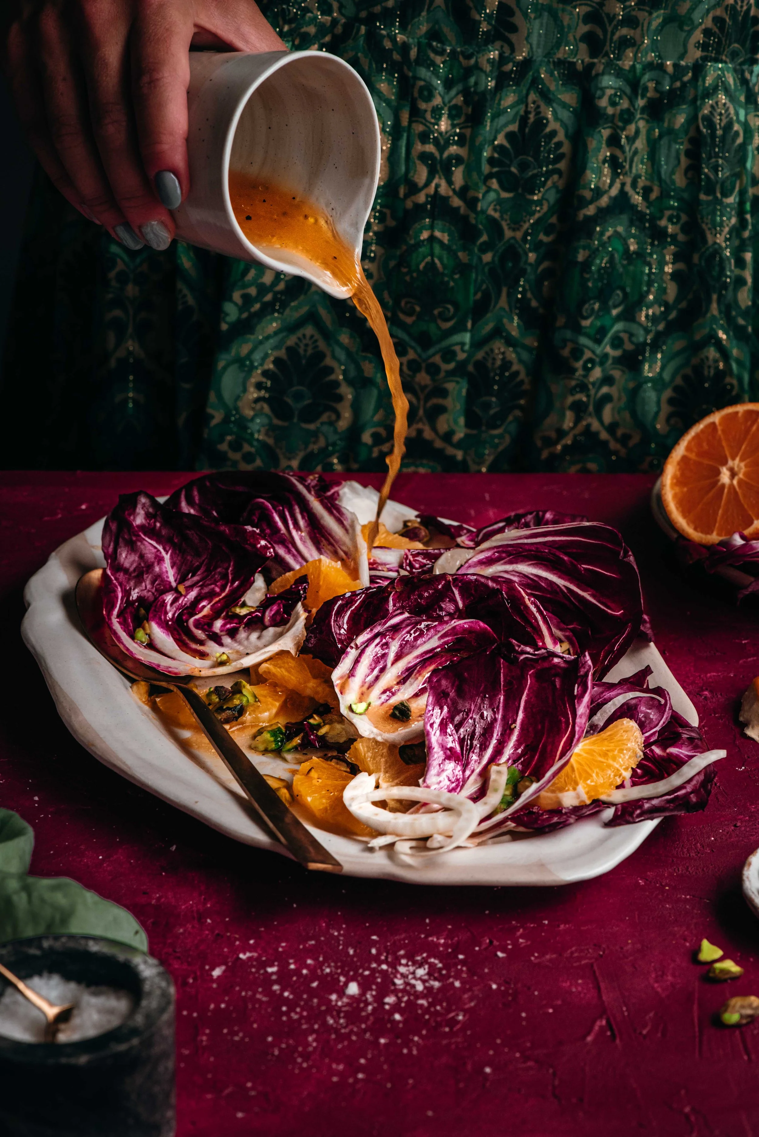 A person pouring a dressing or sauce onto radicchio and citrus salad on a white plate, with a dark red table and sliced oranges nearby.