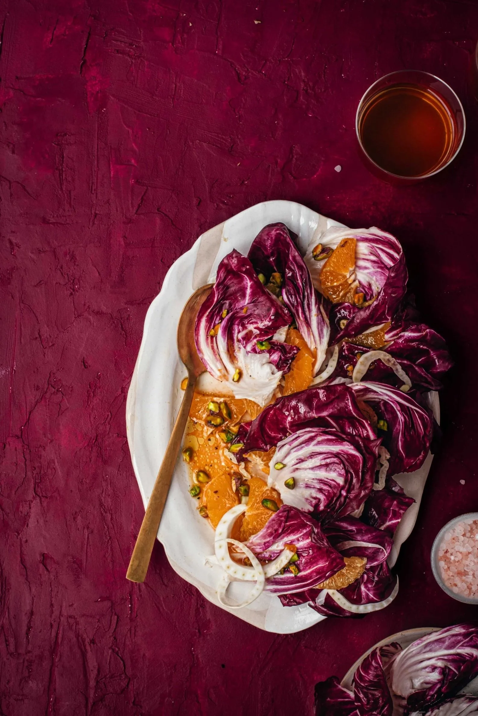 Radicchio and orange salad on a white plate with pistachios, sitting on a dark red textured surface with a glass of dark drink nearby.