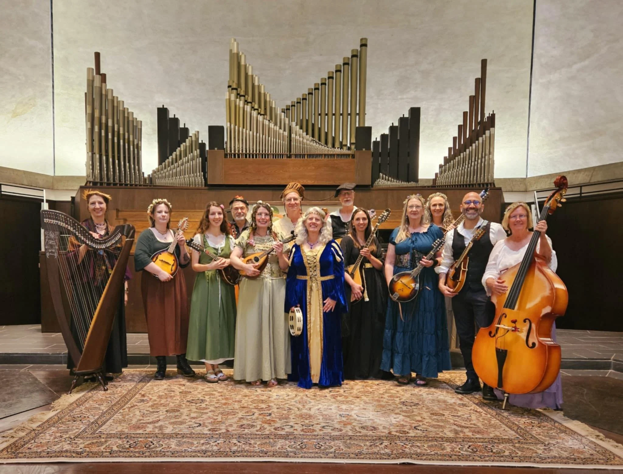 Musicians with mandolins, a harp, and an upright bass stand in front of an organ dressed in medieval costumes