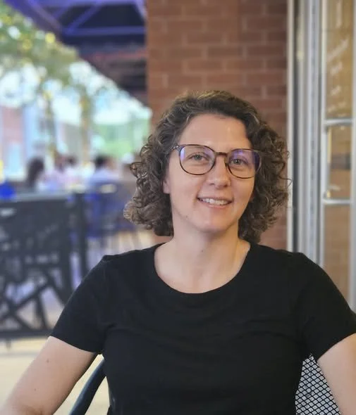 Woman with short, curly brown hair, glasses and a black t-shirt
