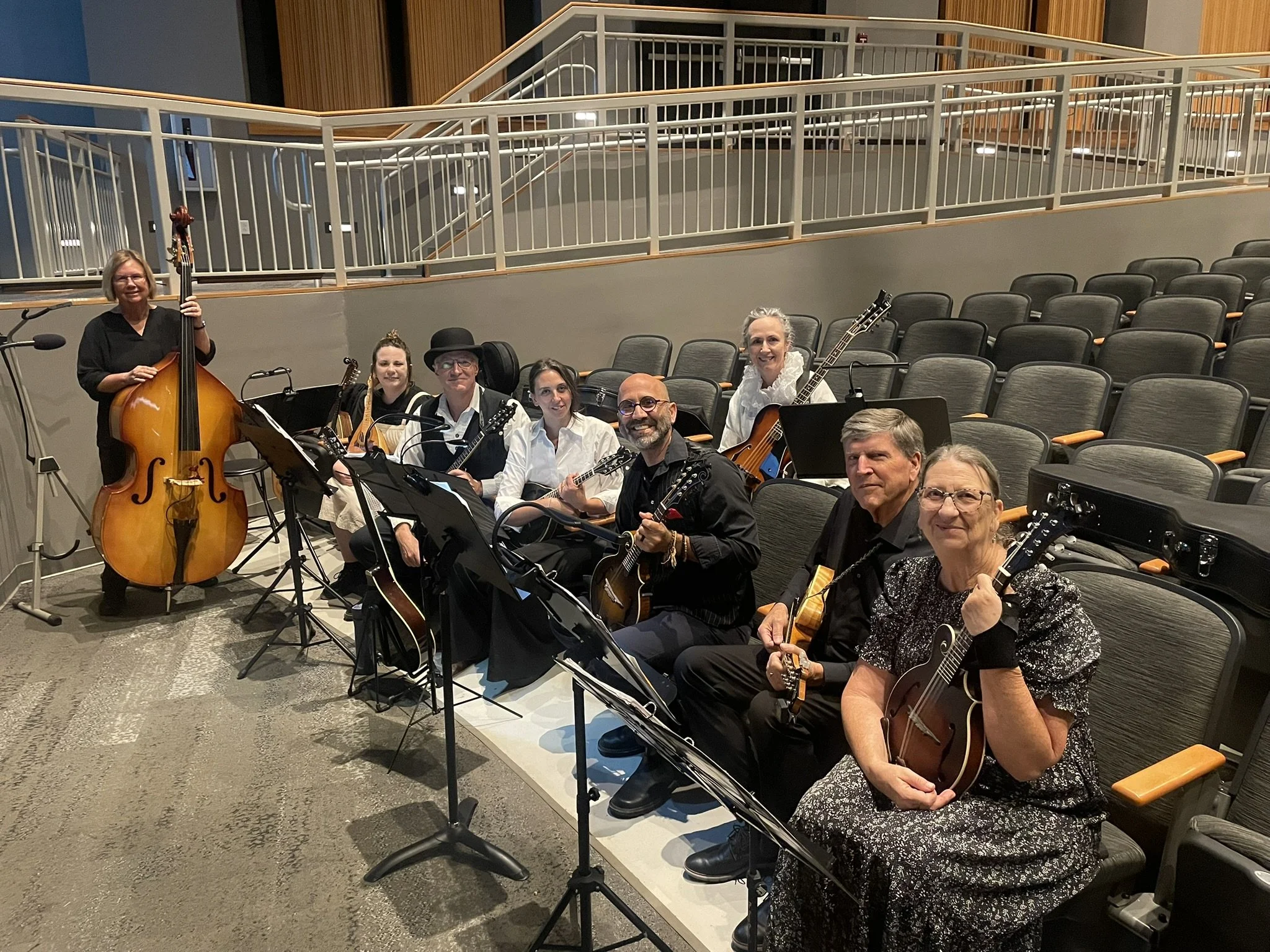 Musicians with mandolins and an upright bass sit in an auditorium smiling at the camera