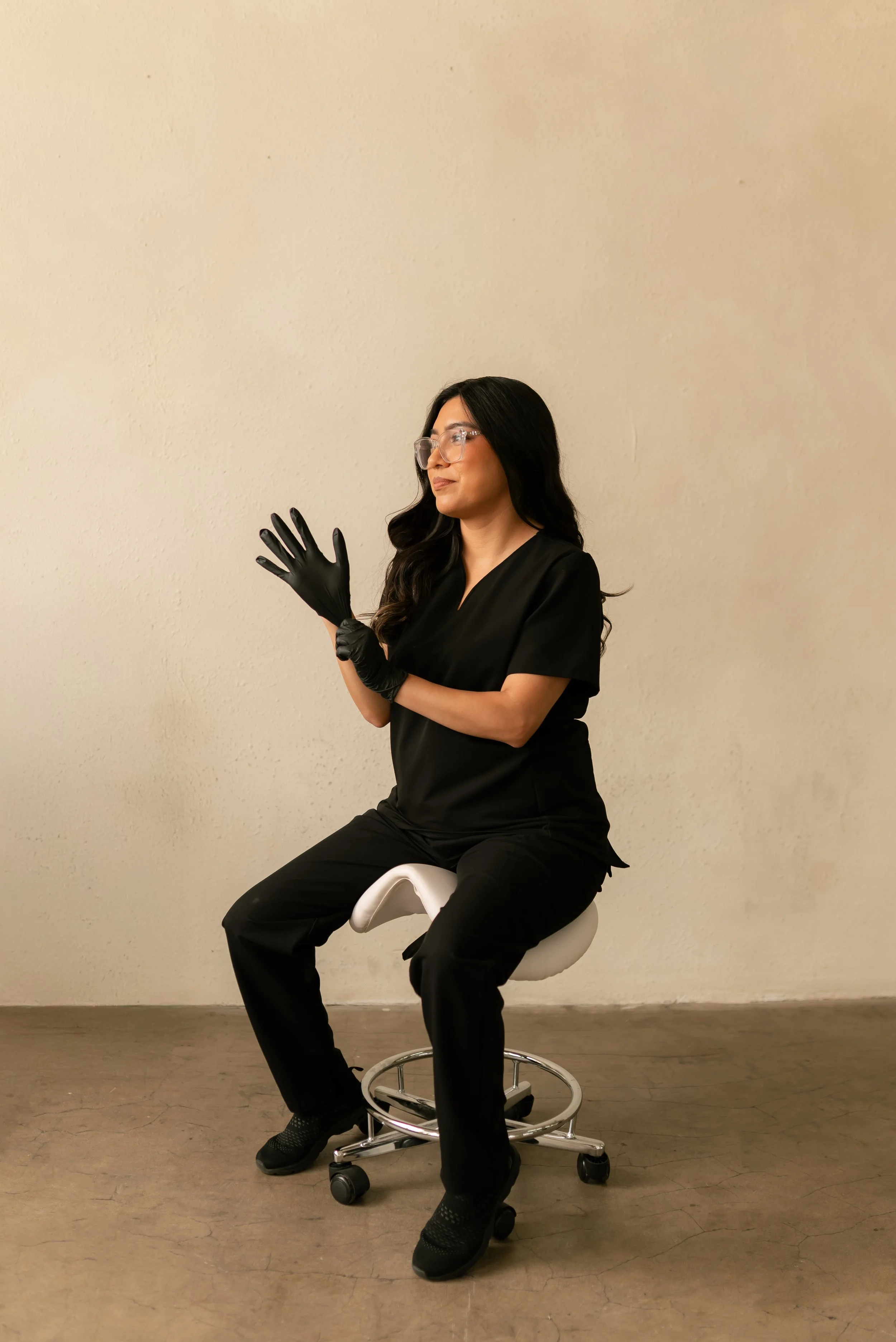 Esthetician in black scrubs putting on gloves before a waxing appointment, showing clean, professional prep in a comfortable studio setting.