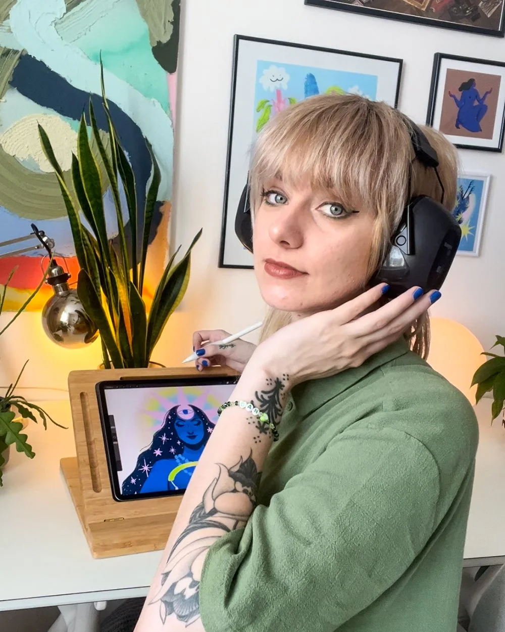 A woman with blonde hair and tattoos is wearing large black headphones, holding her face with one hand, and sitting at a desk with an iPad displaying an artistic illustration of a mystical woman with glowing eyes. She's wearing a green shirt and has blue nail polish.