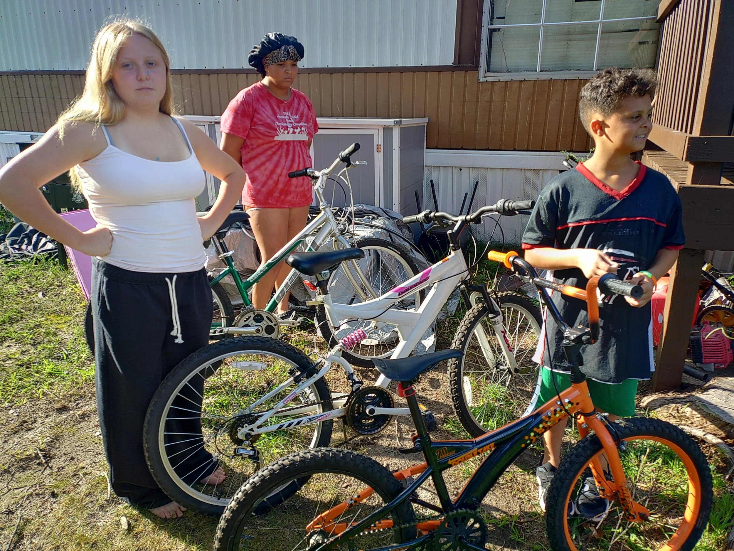 Three children standing outdoors with bikes, in front of a building with metal siding and a wooden fence.