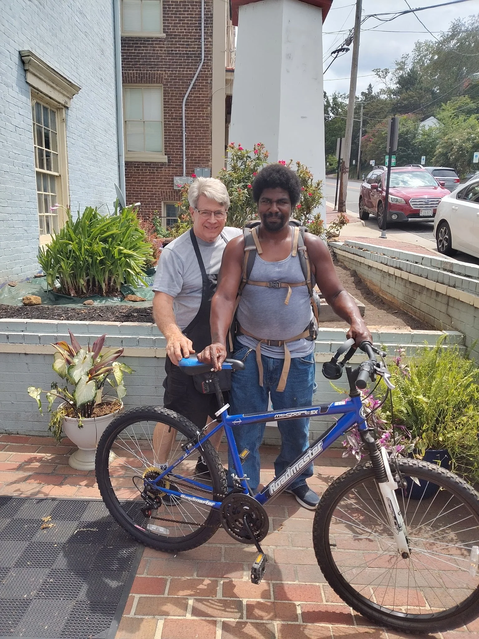 Two men standing outdoors next to a blue mountain bike, with plants, a building, and parked cars in the background.