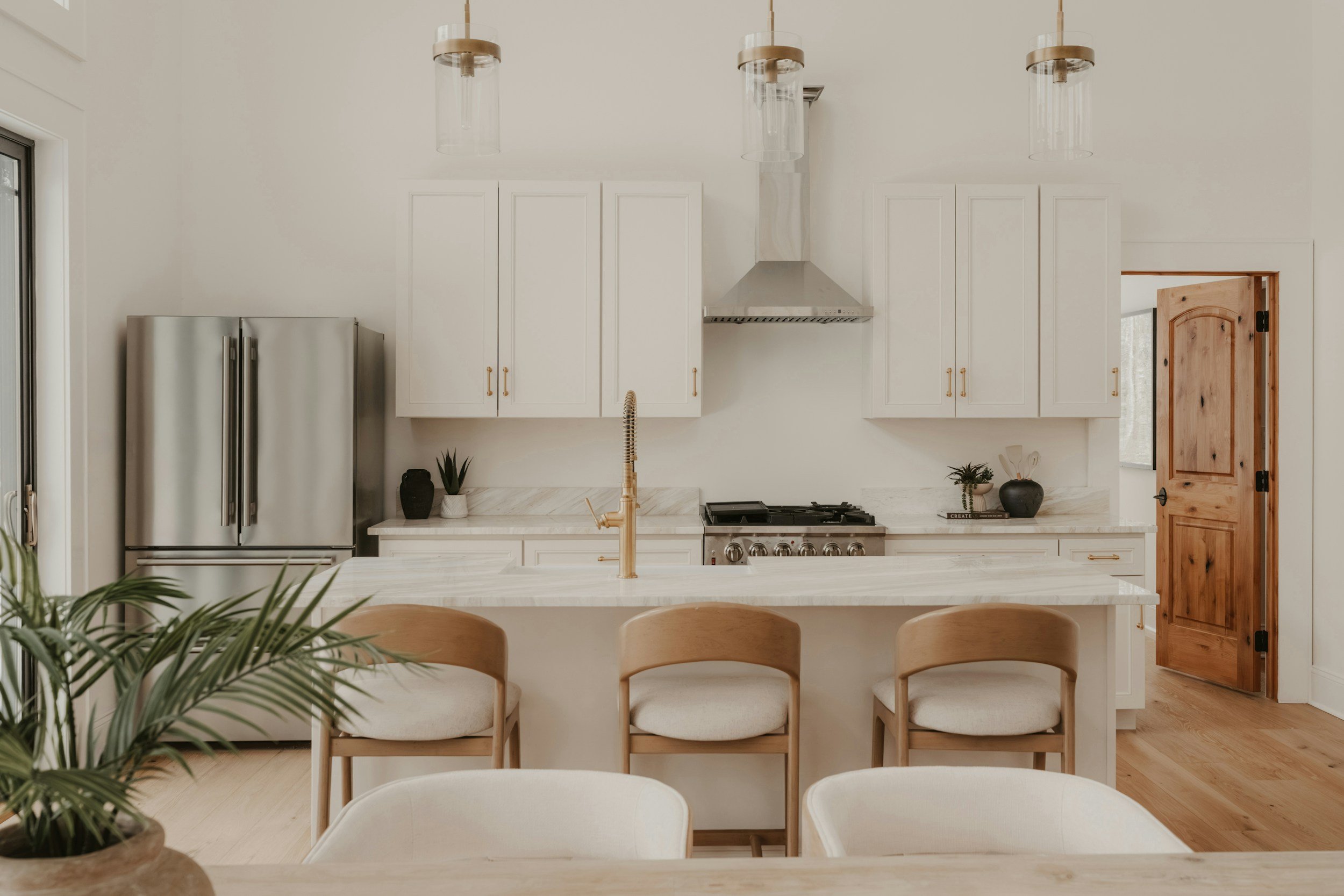 A modern kitchen with white cabinets, stainless steel refrigerator, wooden door, and bar stools around a kitchen island with a marble countertop.