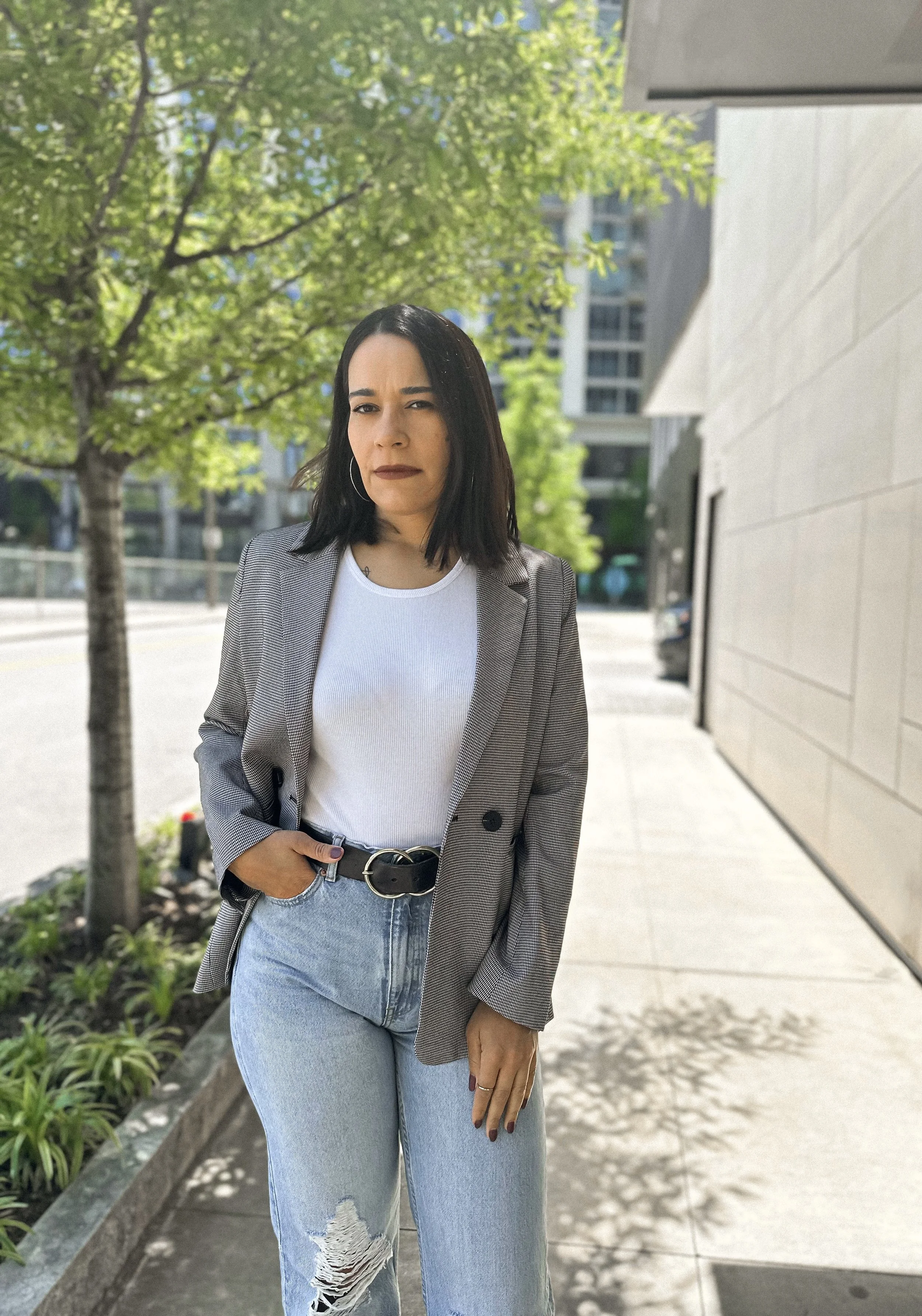 A woman with black hair wearing a gray plaid blazer, white top, and ripped jeans standing outdoors on a sidewalk. Behind her are green trees and modern buildings in an urban setting.