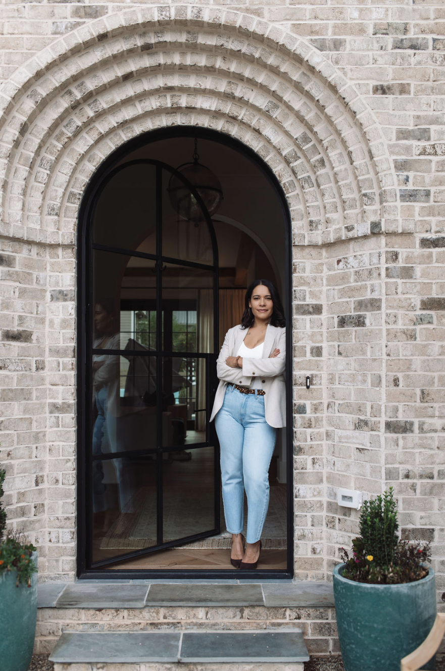 A woman with dark, shoulder-length hair standing at the threshold of a glass-paneled arched door, wearing a white blazer, white top, light blue jeans, and heels, with her arms crossed. The exterior has a brick wall with a potted plant on either side of the steps.