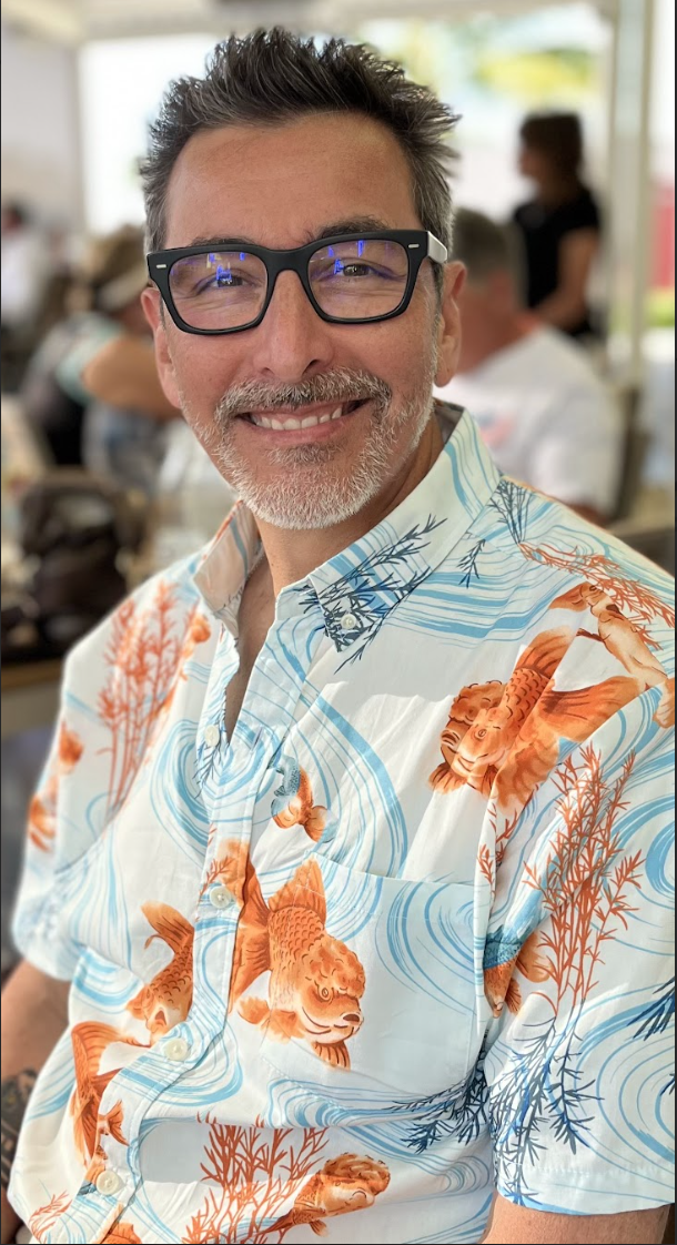 Smiling man with gray beard and black glasses, wearing a white Hawaiian shirt with orange koi fish and blue wave patterns, in a busy social setting.