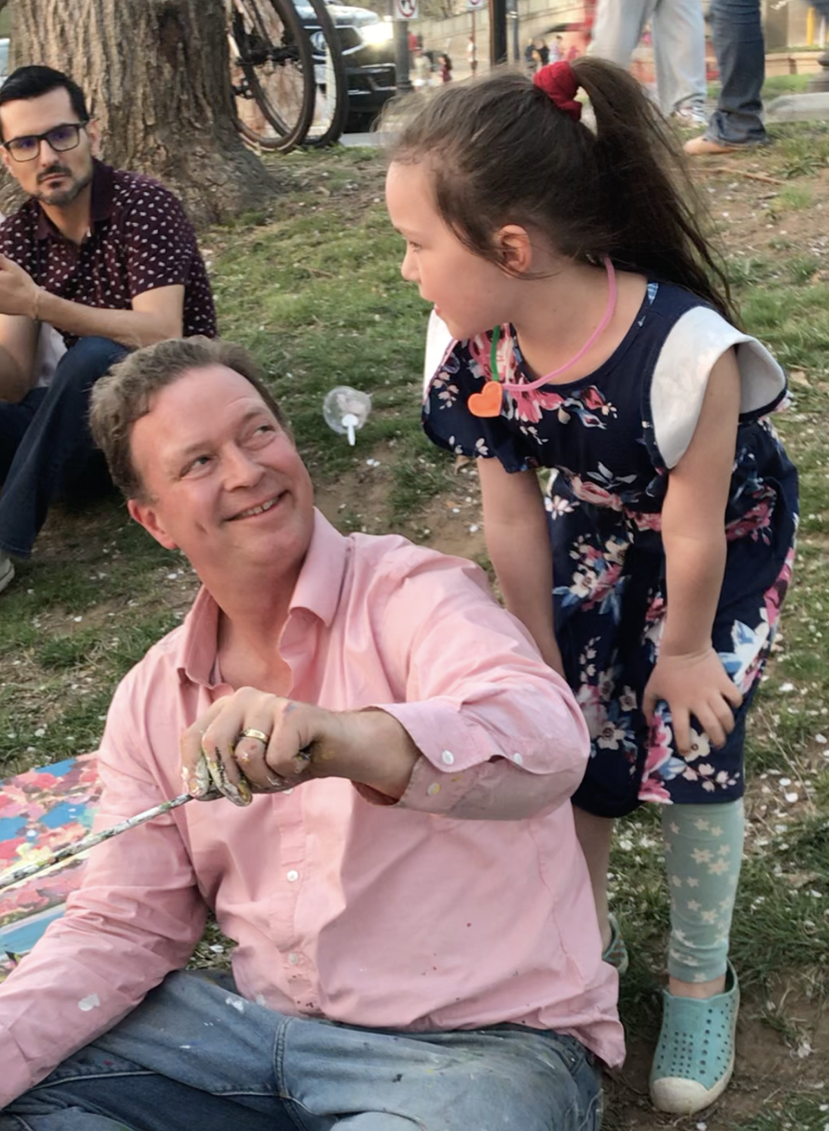 A man with a pink shirt sitting on the ground, smiling at a young girl with a red hair scrunchie, who is leaning over him and smiling. The girl is wearing a navy floral top and leggings with star patterns. Two other people are sitting nearby on the grass, with bicycles and trees in the background.