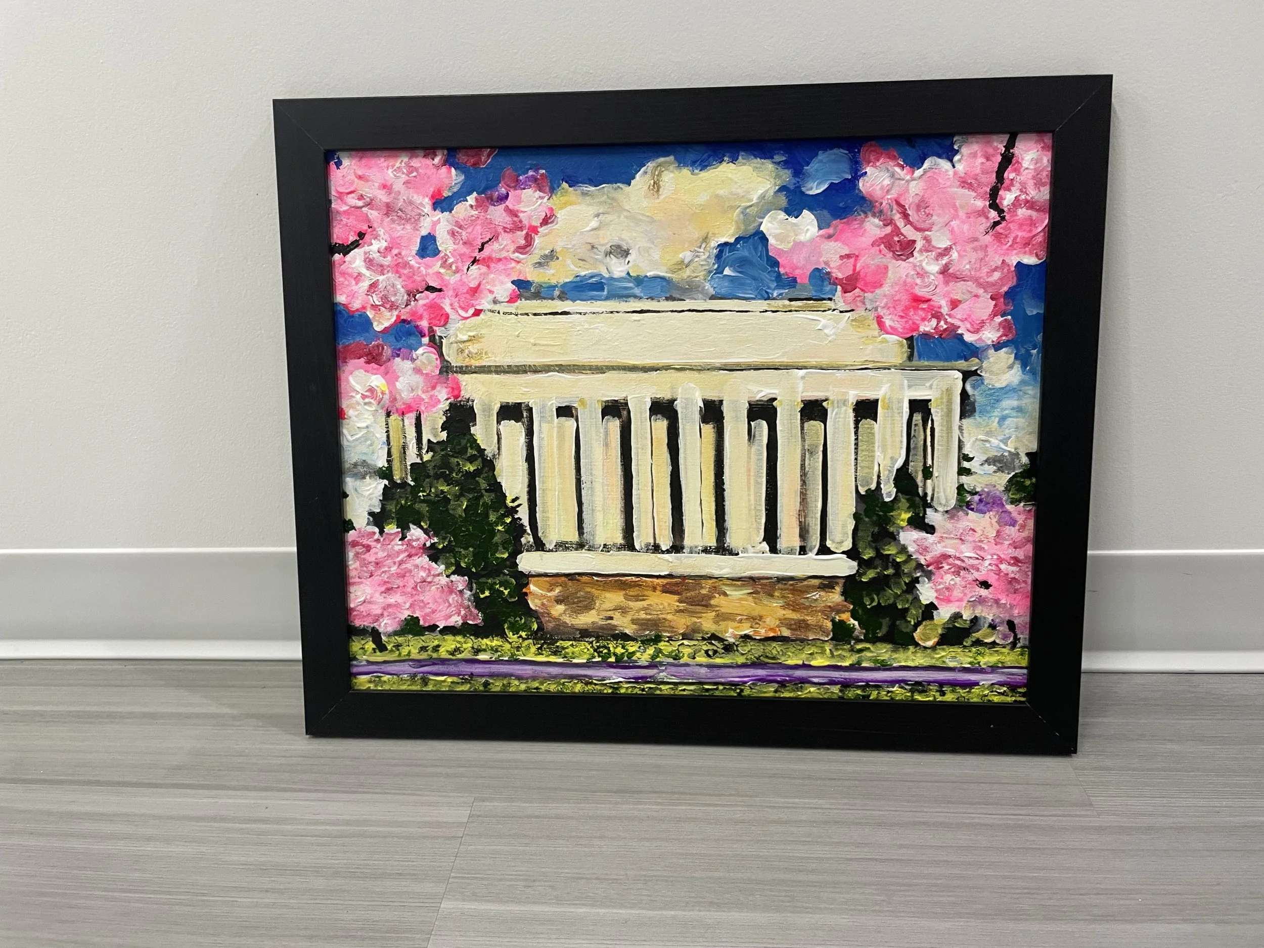 A framed impressionist painting of pink cherry blossoms on a tree with a white fence, blue sky, and clouds in the background, resting on a gray wooden surface.