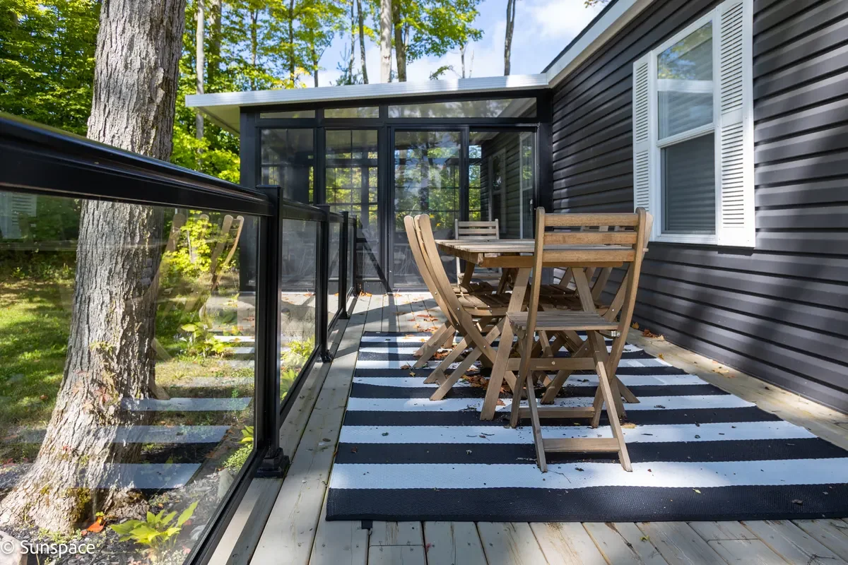 A wooden deck with a black and white striped rug, outdoor wooden furniture, a glass railing, and a screened-in porch attached to a house with gray siding and white shutters, surrounded by trees.
