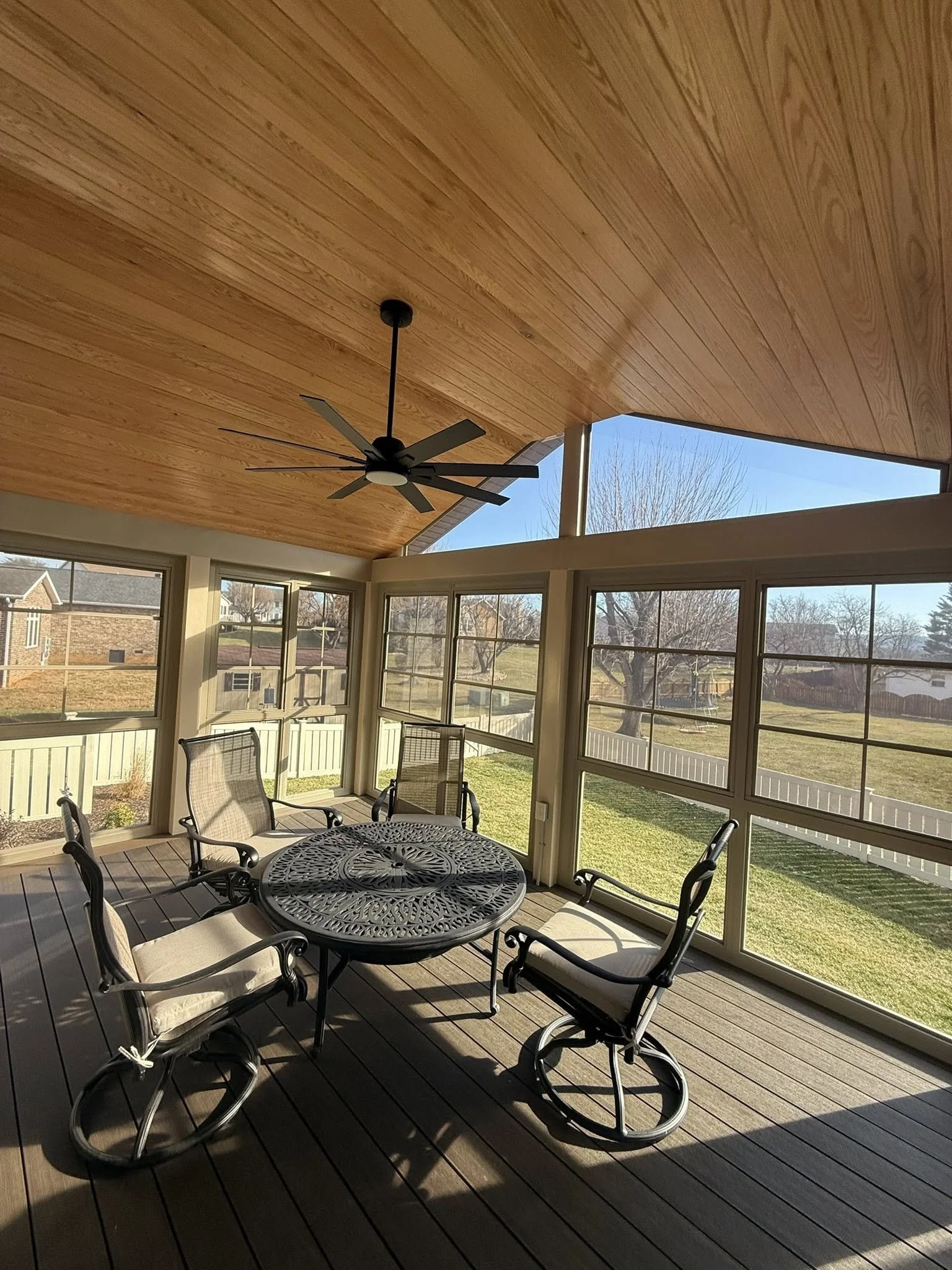 Sunroom with wooden ceiling, ceiling fan, round metal table with decorative pattern, six cushioned chairs, large windows showing backyard with trees, grass, and neighboring houses.