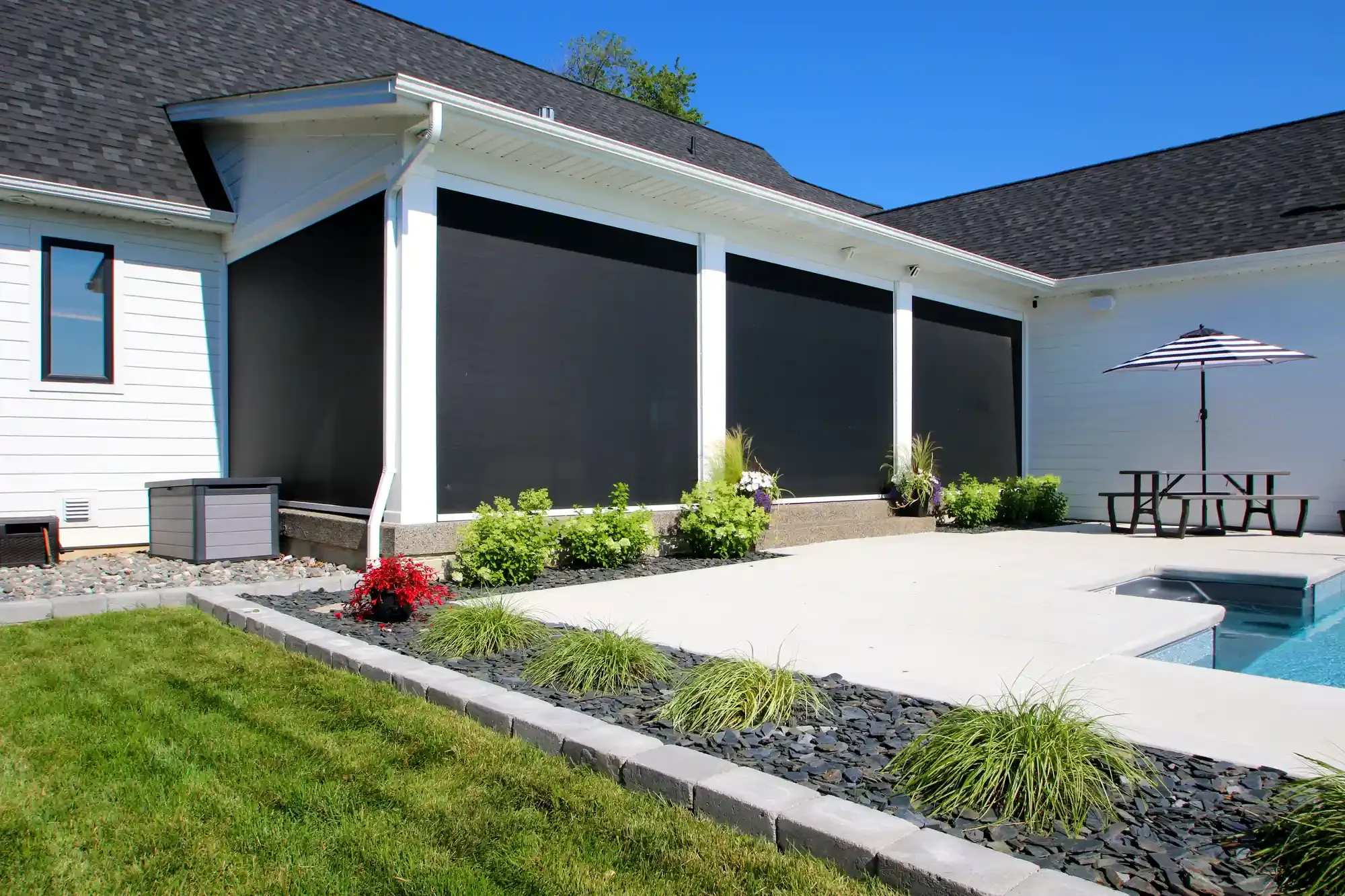 View of a modern house's backyard with a patio, garden, and black screen enclosures, under a clear blue sky.