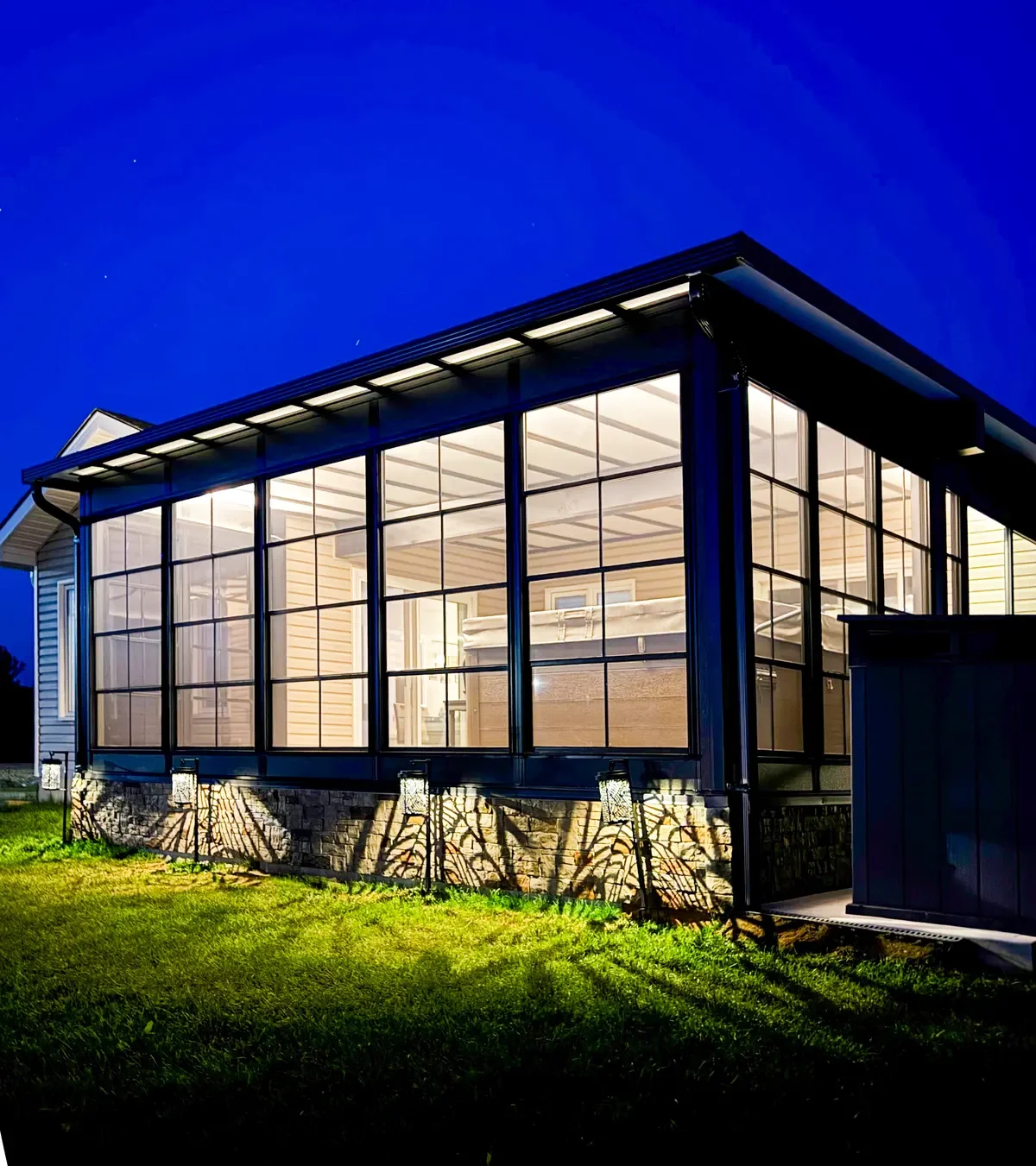 A well-lit screened porch attached to a house at night, featuring large windows, a hot tub inside, and outdoor landscape lighting on the grass.