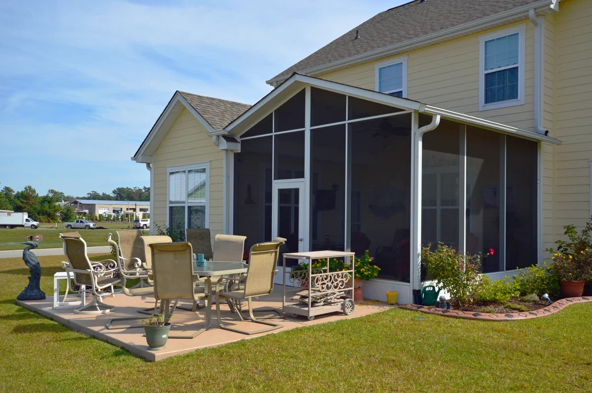 A backyard patio with outdoor furniture, a screened-in porch, and garden plants in a suburban neighborhood.