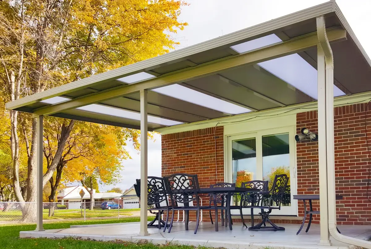 Backyard patio with a metal table and chairs under a covered awning attached to a brick house, with trees displaying fall foliage in the background.