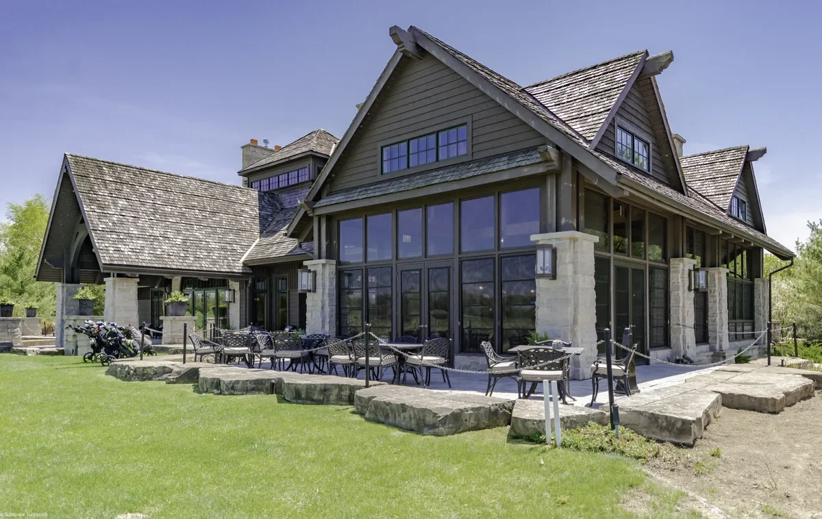 Large modern house with stone pillars and large glass windows, surrounded by a grassy yard with outdoor seating and golf bags.