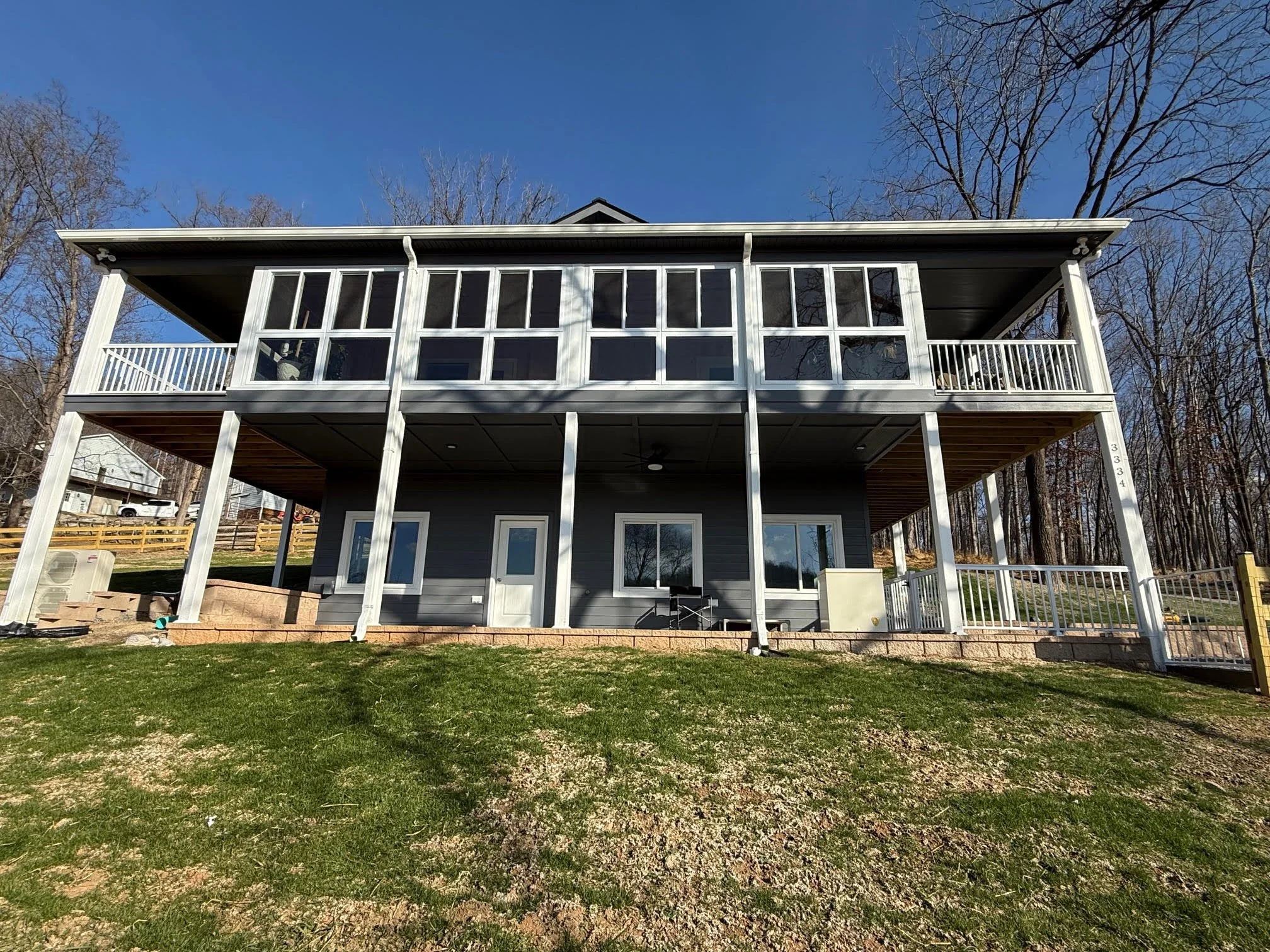 Two-story modern house with a gray exterior, large windows, and a wraparound balcony on the upper floor. The house is situated on a grassy hillside with a wooded background and a clear blue sky.