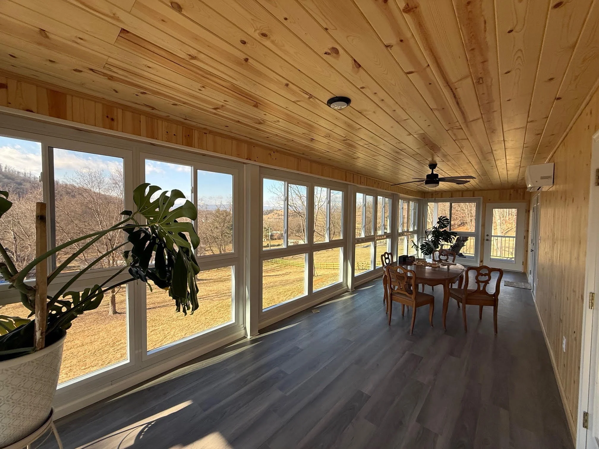 A sunlit enclosed porch with large windows overlooking a grassy backyard and distant hills. Inside, there's a round wooden dining table with five ornate wooden chairs, potted plants, a ceiling fan, and mounted air conditioning unit. The ceiling and w