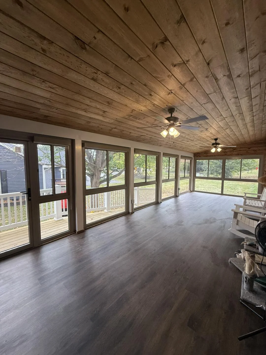 Empty screened porch with wooden ceiling, large windows, ceiling fans, and outdoor chairs.