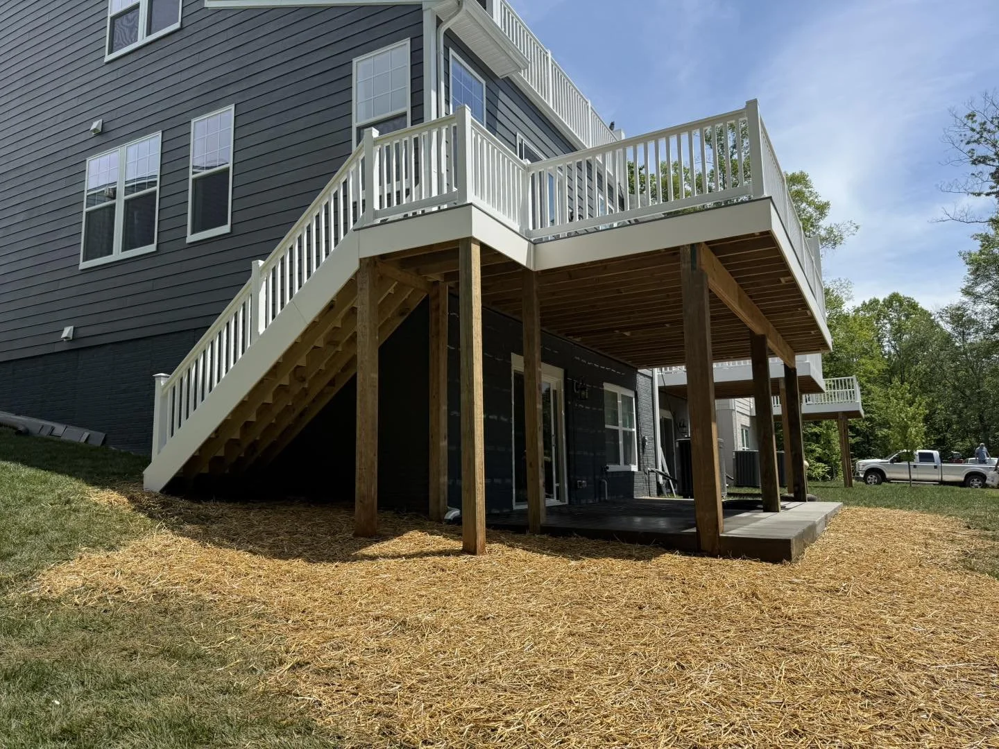 Back of a modern house with a large elevated deck and stairs, supported by wooden beams, with black siding and multiple windows, on a sunny day with a blue sky and green trees in the background.