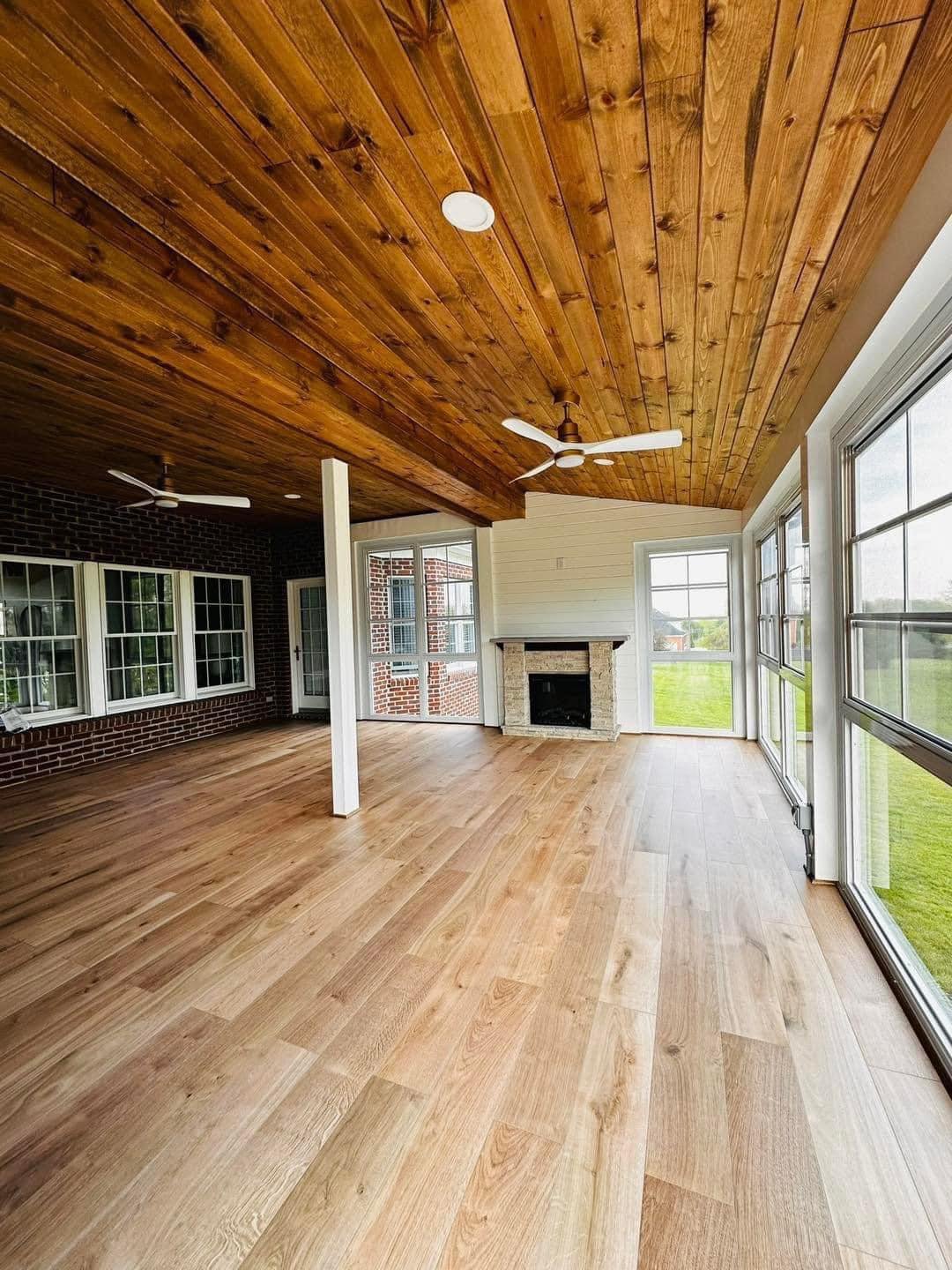 Bright living room with wood-paneled ceiling, hardwood flooring, fireplace, ceiling fans, and large windows overlooking a grassy outdoor area.