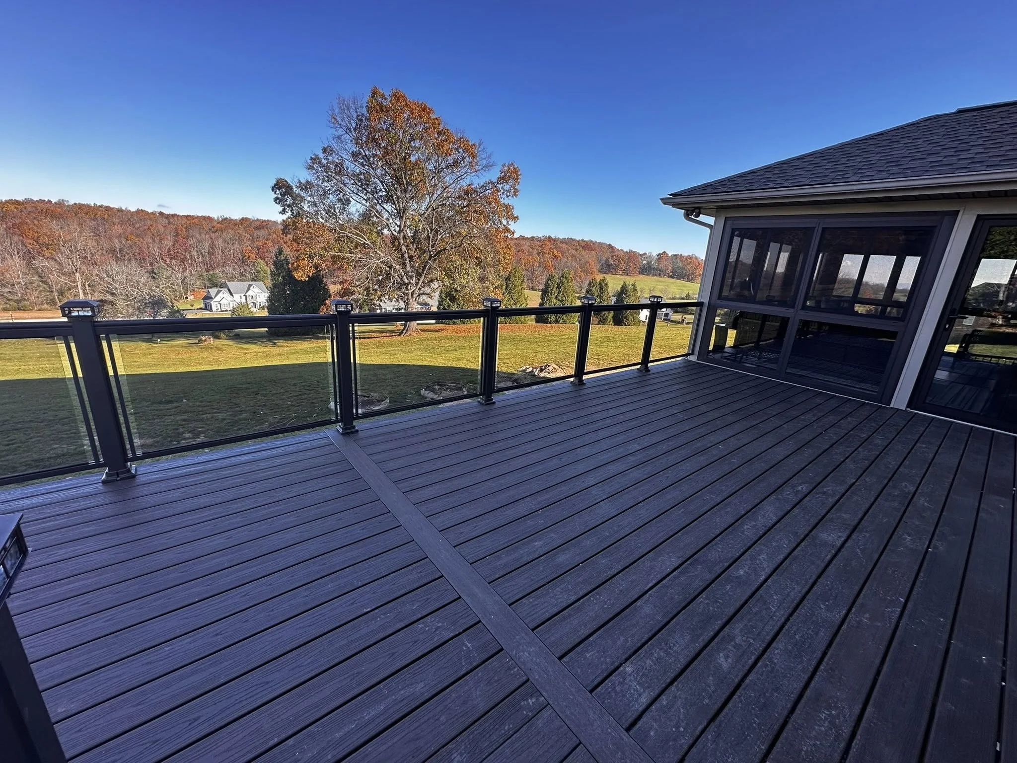 A wooden deck with glass railings overlooks a field with trees displaying fall foliage and a quiet neighborhood with houses in the distance under a clear blue sky.