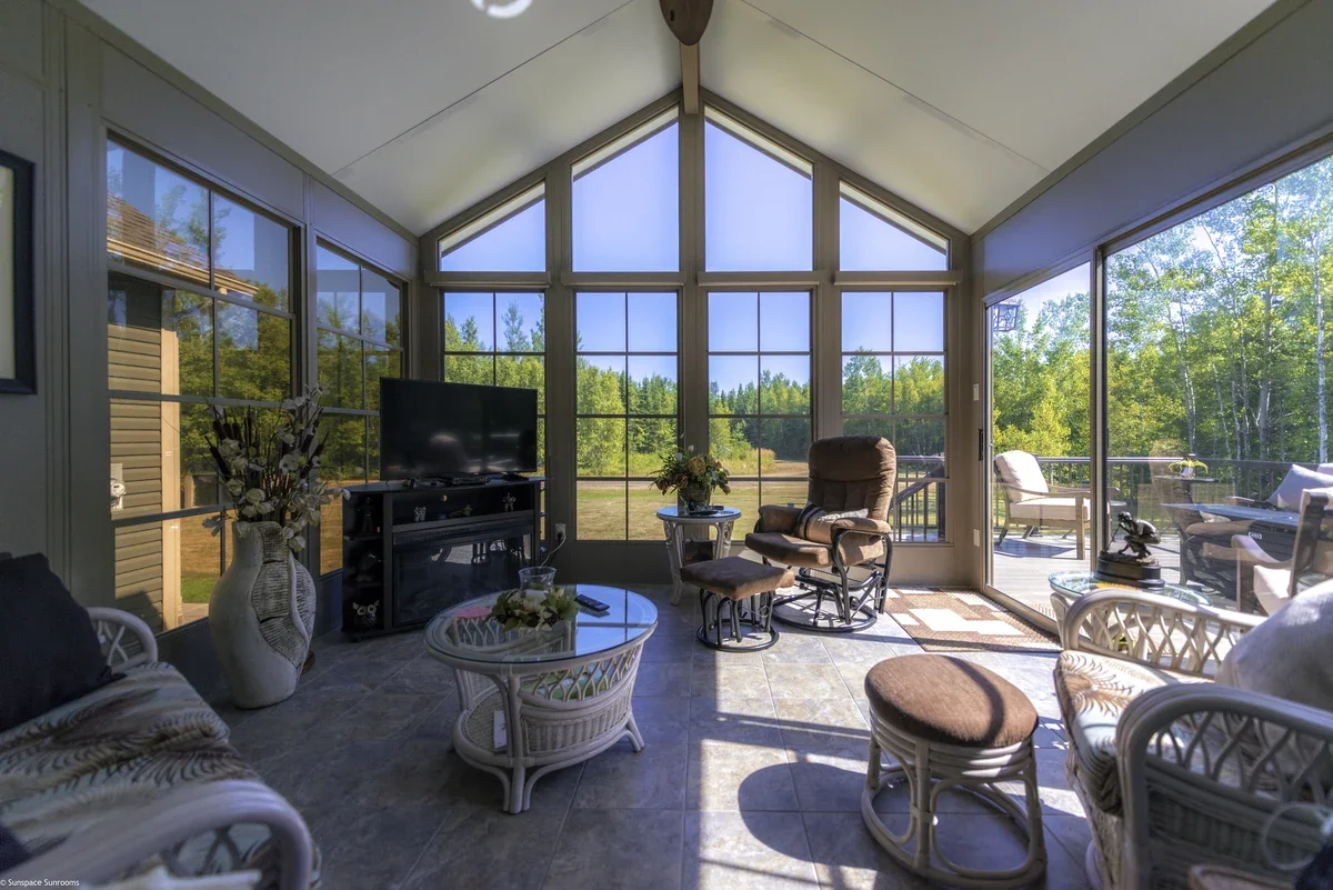 Sunroom with large windows showing a green outdoor landscape, furnished with chairs, a small table with a floral arrangement, and a television.