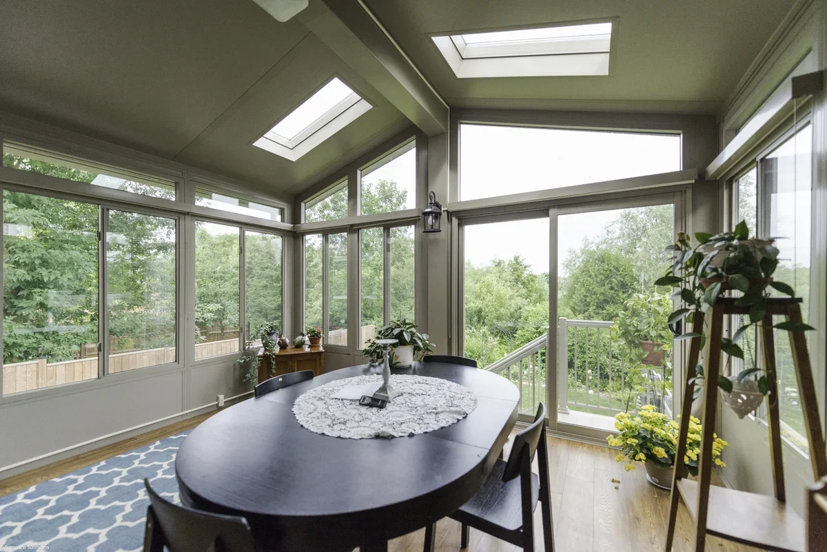 Sunroom with large windows, skylights, and wooden flooring, furnished with a black dining table, chairs, potted plants, and a patterned rug.