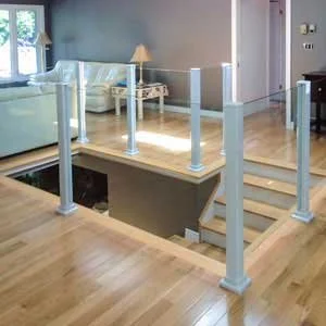 Interior view of a house with a staircase surrounded by a glass railing, a living room area with a white sofa, side table, and lamp.