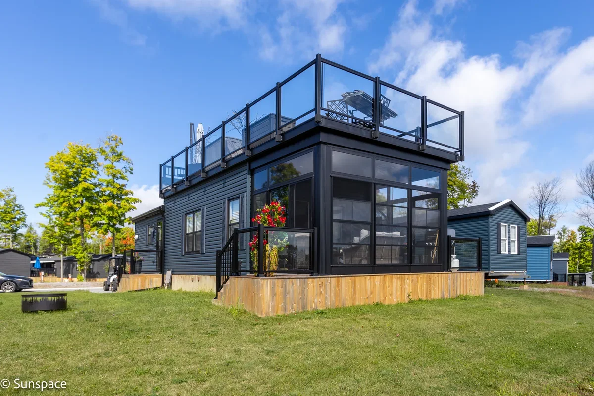 A two-story modern house with a black metal exterior and large glass windows, featuring a rooftop deck with outdoor furniture, a small porch with potted plants, and surrounded by a grassy yard with trees and other similar houses in the background.
