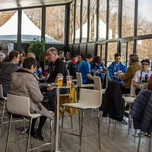 People sitting and socializing inside a bright, glass-enclosed venue with large windows and trees outside.