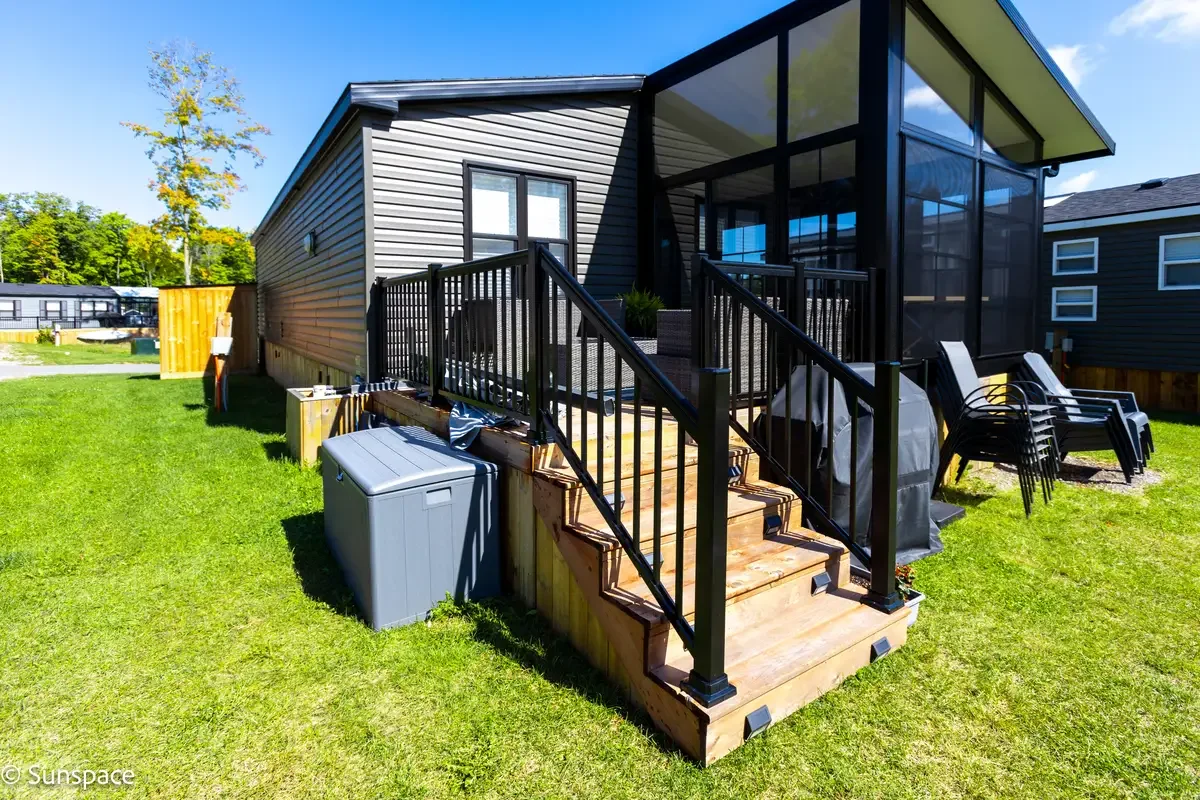Backyard with a screened porch, wooden stairs with black railing, lawn, patio chairs, and a storage box, in a sunny suburban neighborhood.