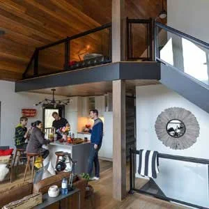 A modern open-concept kitchen and living area with exposed wooden ceiling and a staircase leading to an upper level, with four people socializing around a kitchen island.