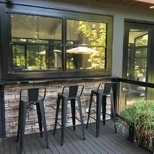Indoor seating area with four black bar stools along a counter, overlooking a patio with trees and an umbrella outside.