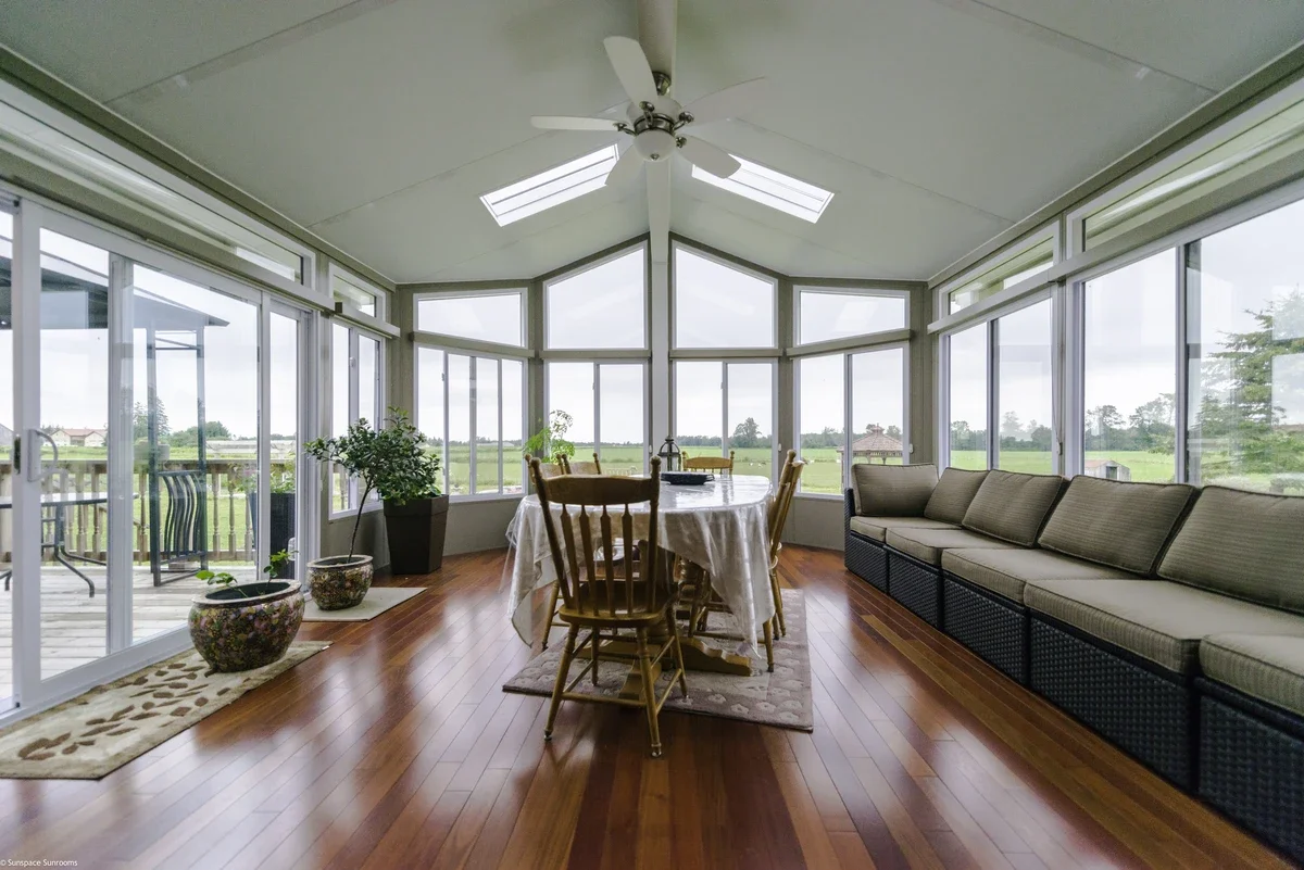 Sunroom with large windows, wooden flooring, a dining table with chairs, potted plants, and a sofa with cushions.