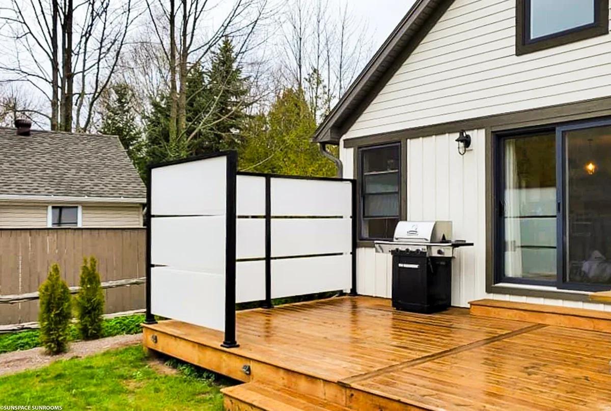 A backyard deck with a grill, a privacy screen, and sliding glass door leading into a house. The deck is made of wood and has a small step down to the grass. There is a house with white siding and black trim, and trees in the background.