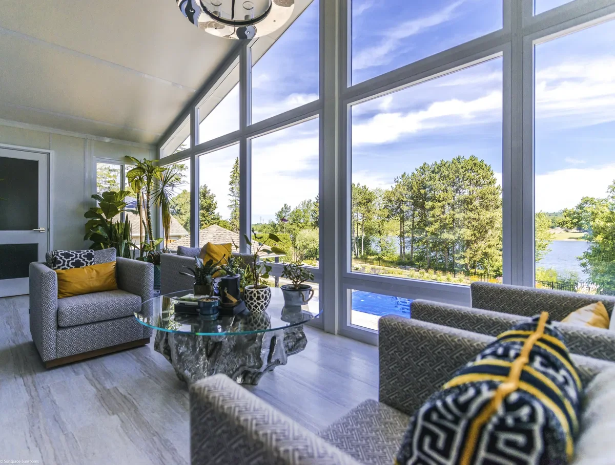 Living room with large floor-to-ceiling windows overlooking a lake and trees, decorated with patterned sofas, yellow and black pillows, and potted plants, and a modern chandelier.