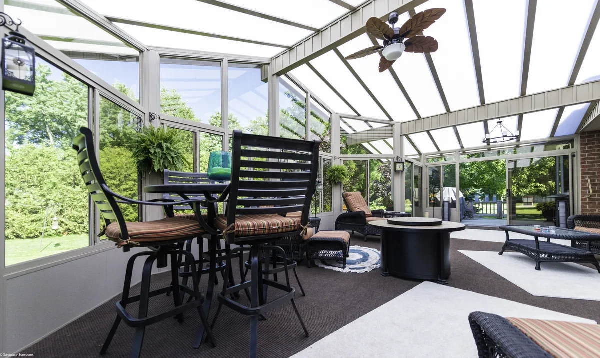 Sunroom with large windows, ceiling fan, wicker furniture, potted plants, and outdoor view of trees and a backyard.