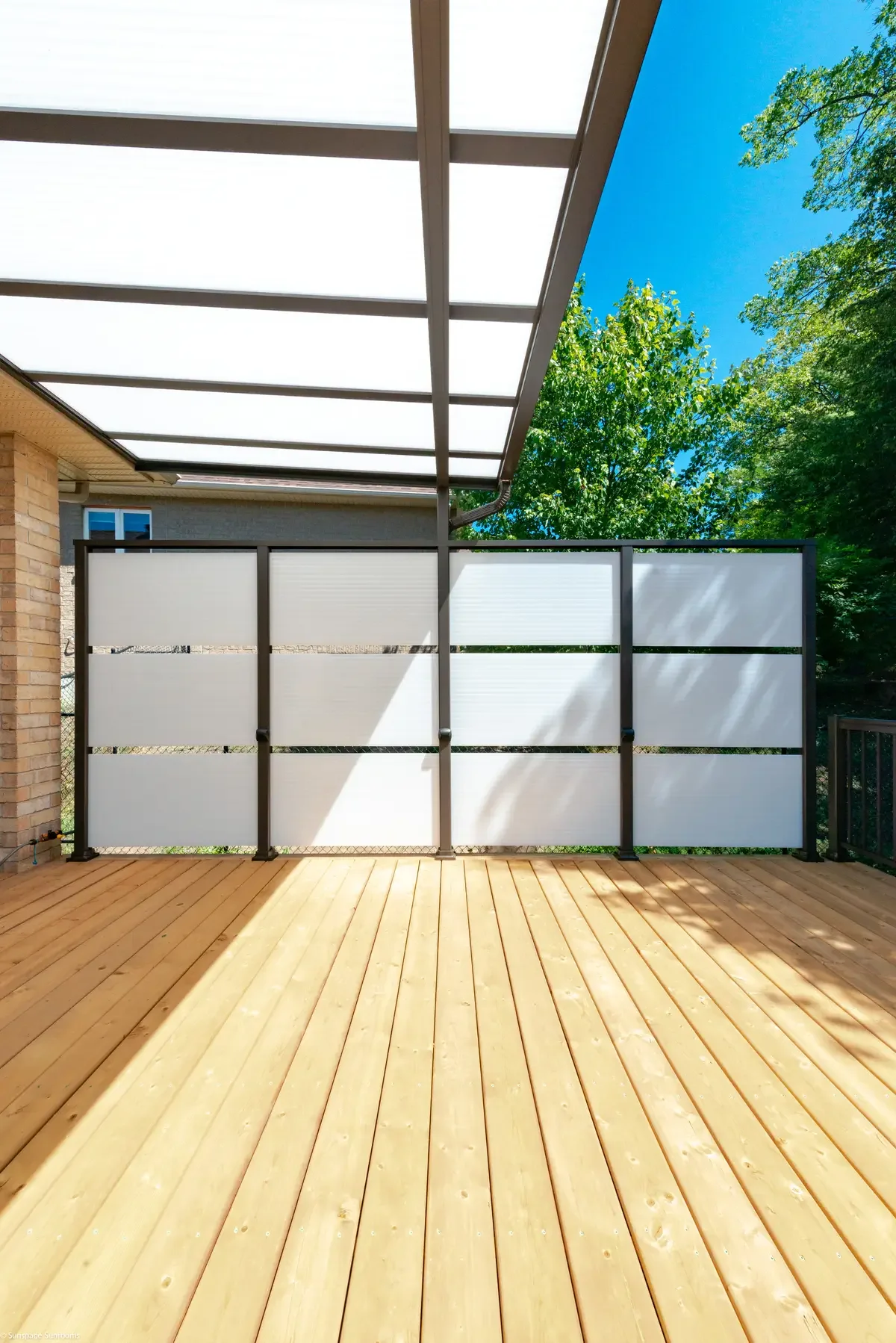 A wooden deck with a white privacy screen, a shaded outdoor area, and green trees and blue sky in the background.