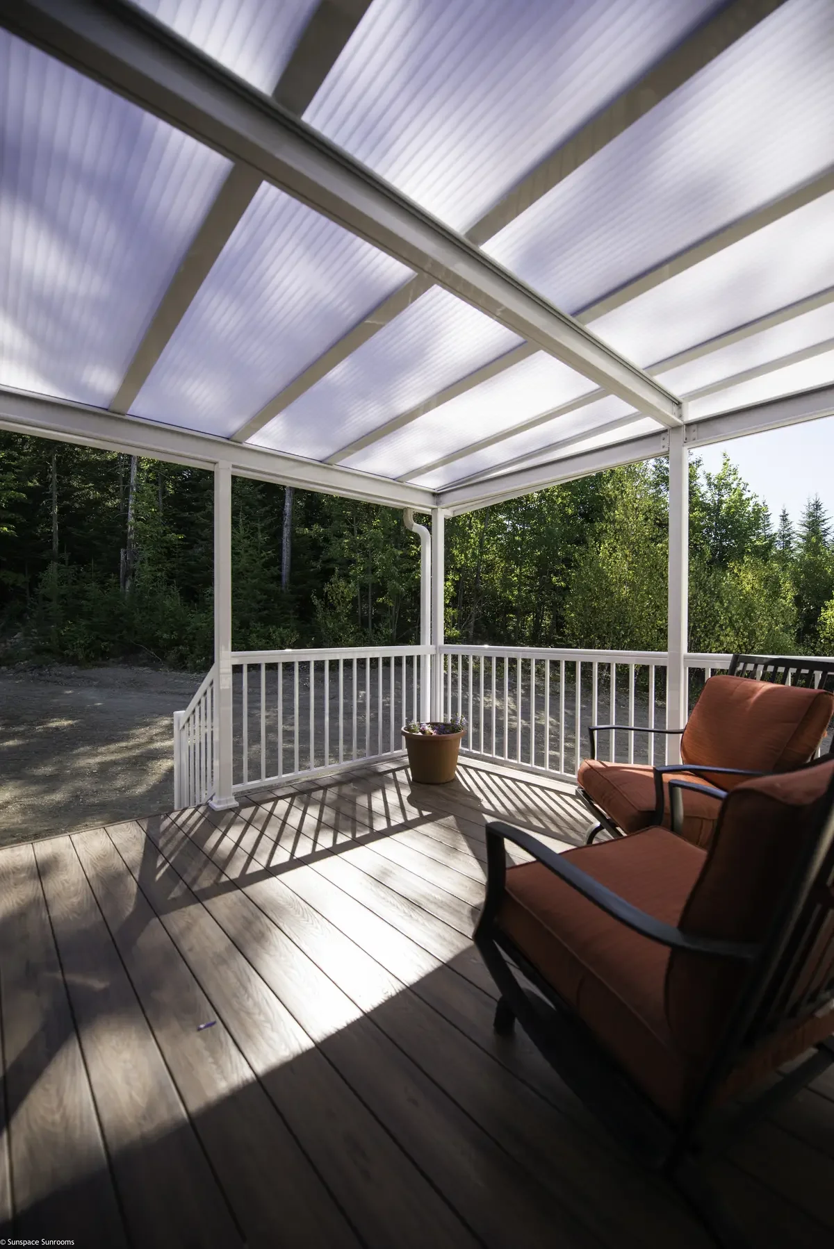 Wooden porch with outdoor chairs, a potted plant, and a white railing, overlooking a wooded area, with a roof overhead casting shadows.