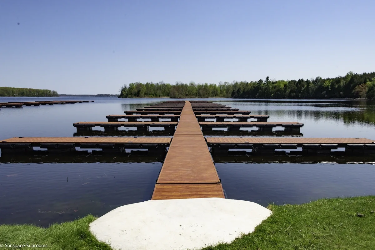 A wooden dock extends into a calm lake, with multiple floating platforms on either side, surrounded by green trees under a clear sky.