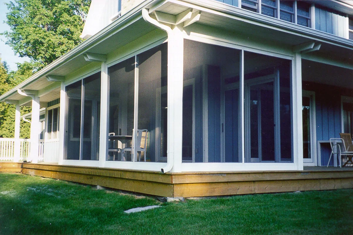 A house with a screened-in porch with sliding glass doors and a wooden deck, surrounded by green grass and trees.