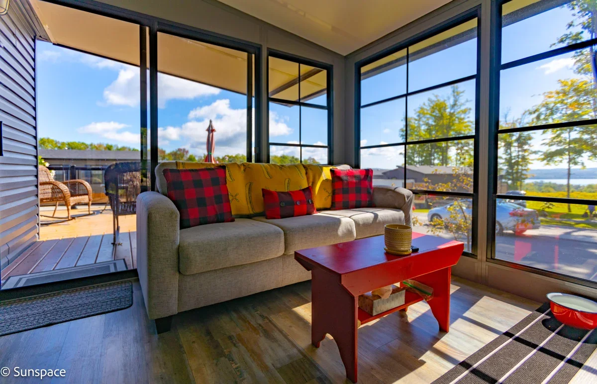Sunroom with large windows showing an outdoor patio and landscape, beige sofa with red and black plaid pillows, a red coffee table with a candle, and woven chairs on the patio.