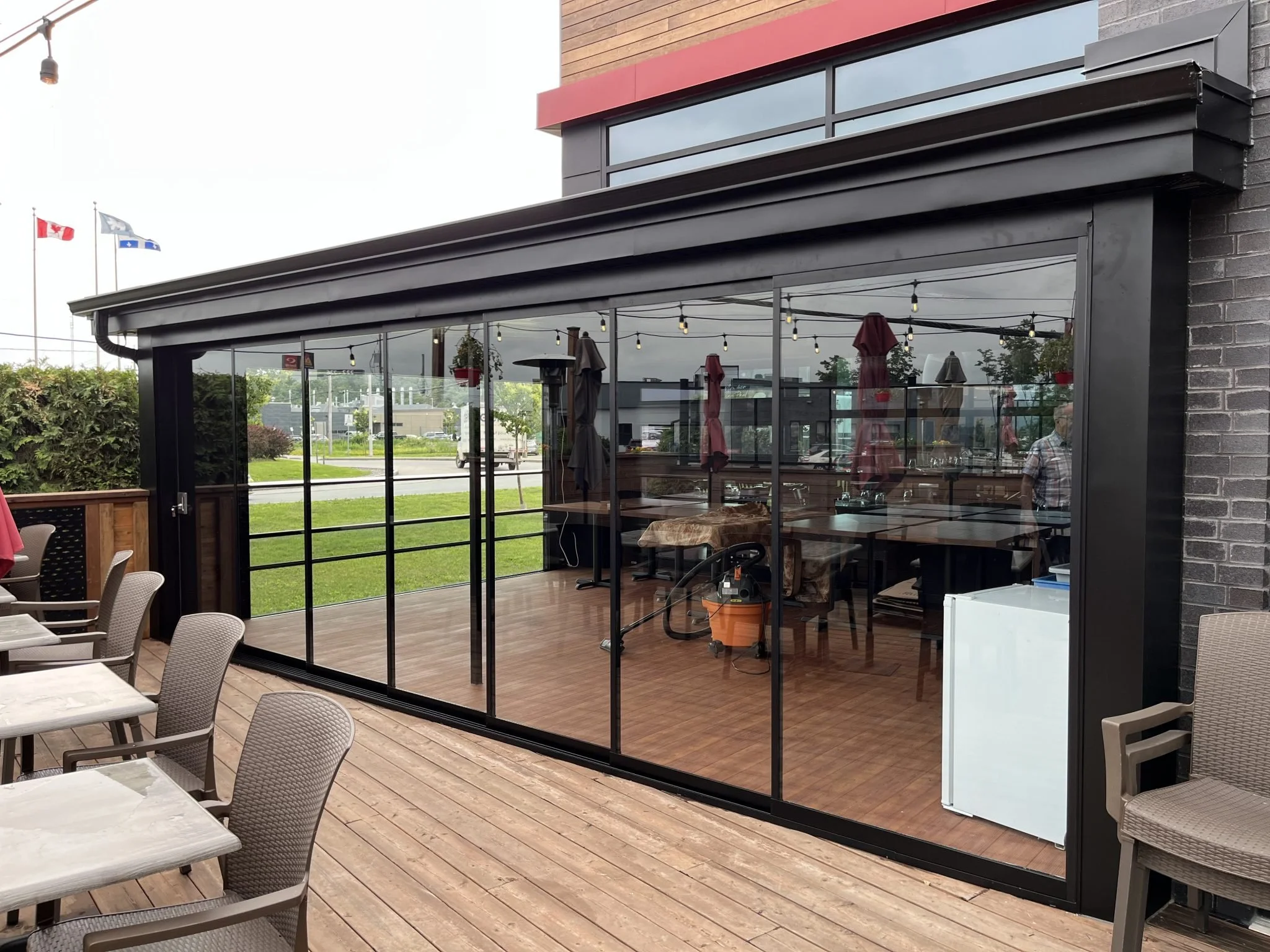 Exterior of a restaurant with large glass windows, a red brick and wood façade, and patio seating with indoor space visible through the windows. Flags are visible in the background.