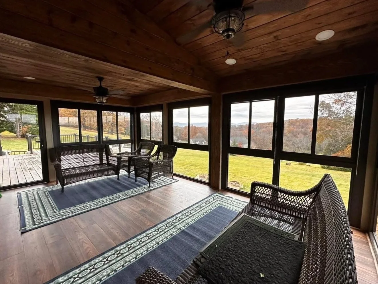 Sunroom with wicker furniture, large windows, wooden ceiling, ceiling fans, and outdoor views of a grassy yard and rolling hills.