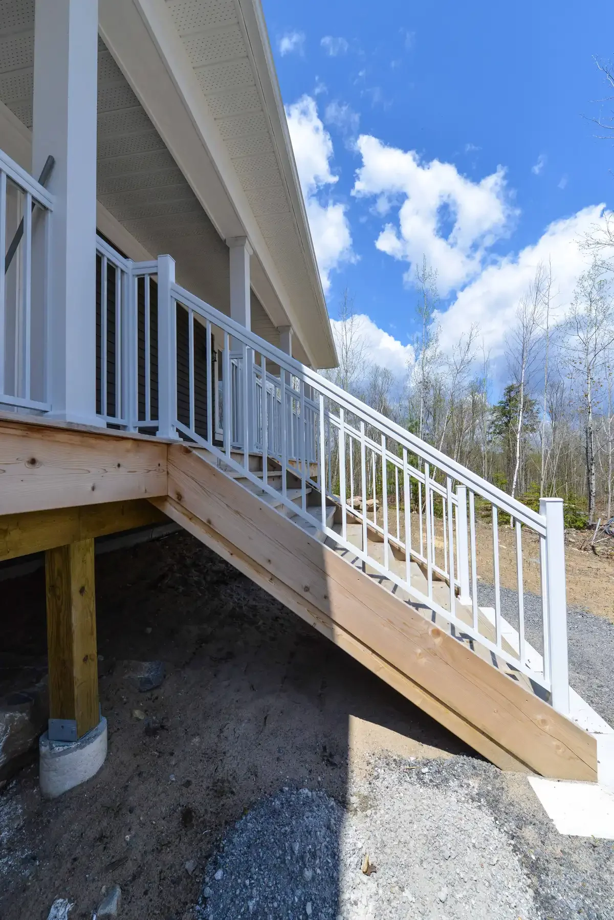 Newly built front porch staircase with white metal railing and wooden supports, attached to a house with light-colored siding, against a background of a partly cloudy sky and leafless trees.