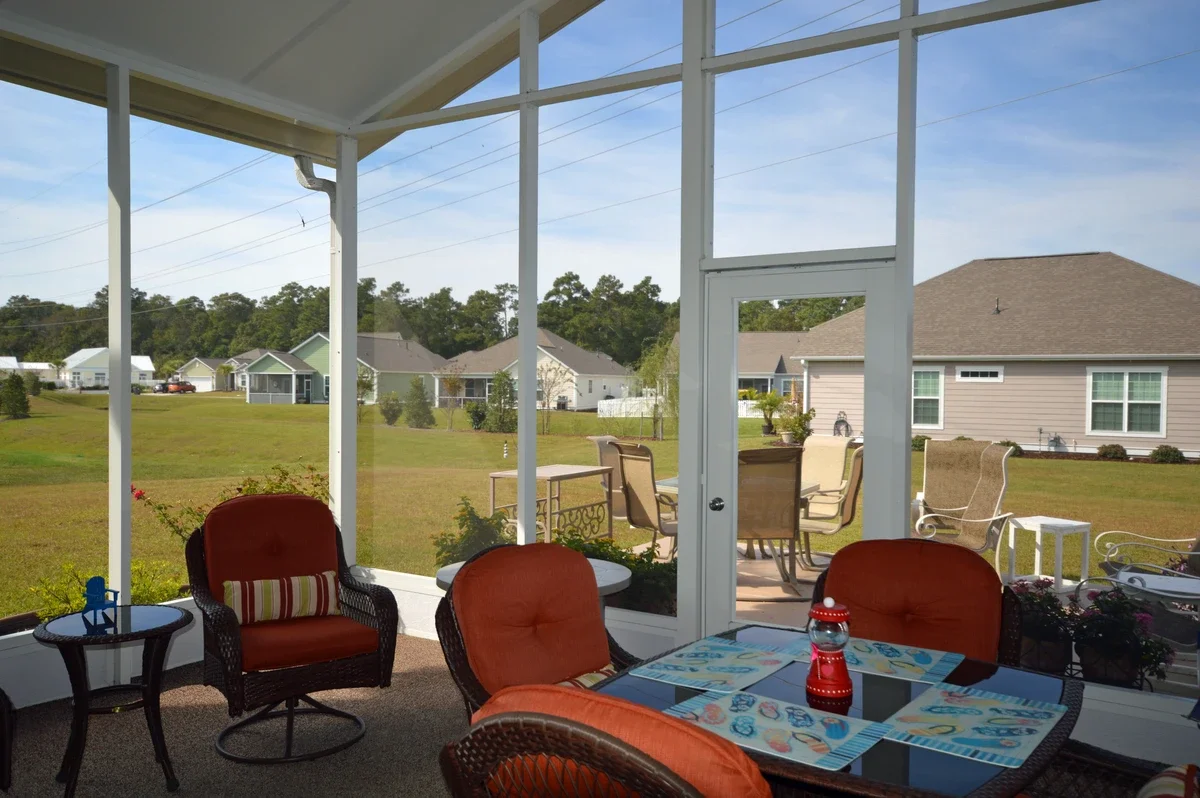 View of a screened porch with outdoor furniture, including a glass-top table with placemats and a decorative red lantern, overlooking a backyard with lawn, patio furniture, neighboring houses, and a partly cloudy sky.